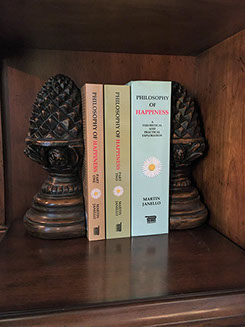 Photo of hardcover book (light blue cover) and the 2-part paperback (green, brown covers) of Philosophy of Happiness book on wooden bookshelf.
