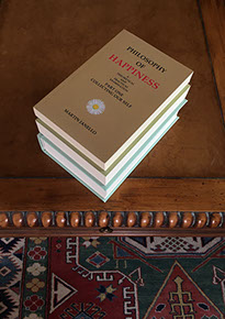 Photo of the hardcover and the 2-part paperback of the Philosophy of Happiness book, diagonally stacked on desk.